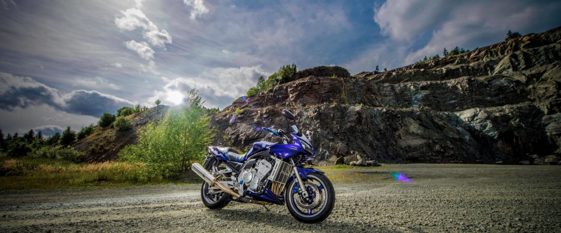 Werbeaufnahme, blaues Motorrad steht auf einem Kiesweg vor einem felsigen, bewaldeten Hintergrund.